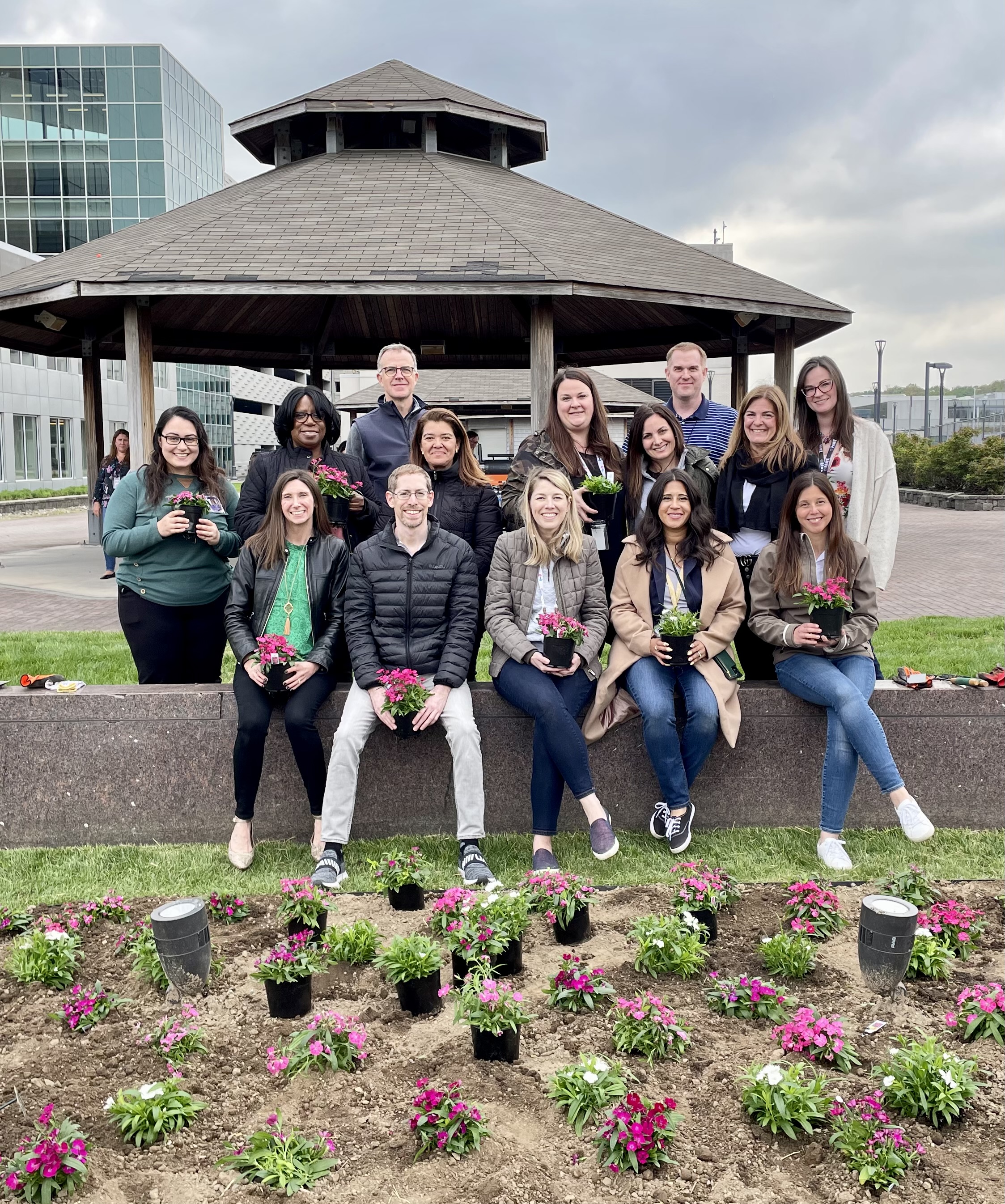 A diverse group of employees holding pots of pink flowers in front of a garden 