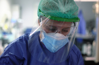 A woman wearing medical scrubs, surgical mask, hair net, safety goggles, and a face shield