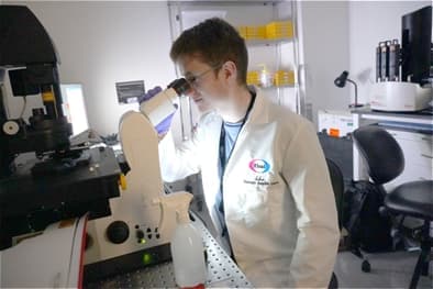 A male scientist wearing a white lab coat and a regular pair of glasses, looking into a microscope