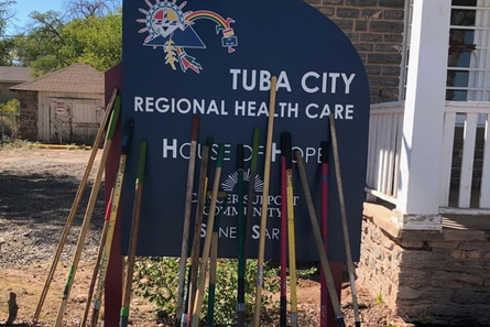 A sign outside a building with text saying Tuba City, Regional Health Care, House of Hope. Wooden sticks lean agains the sign