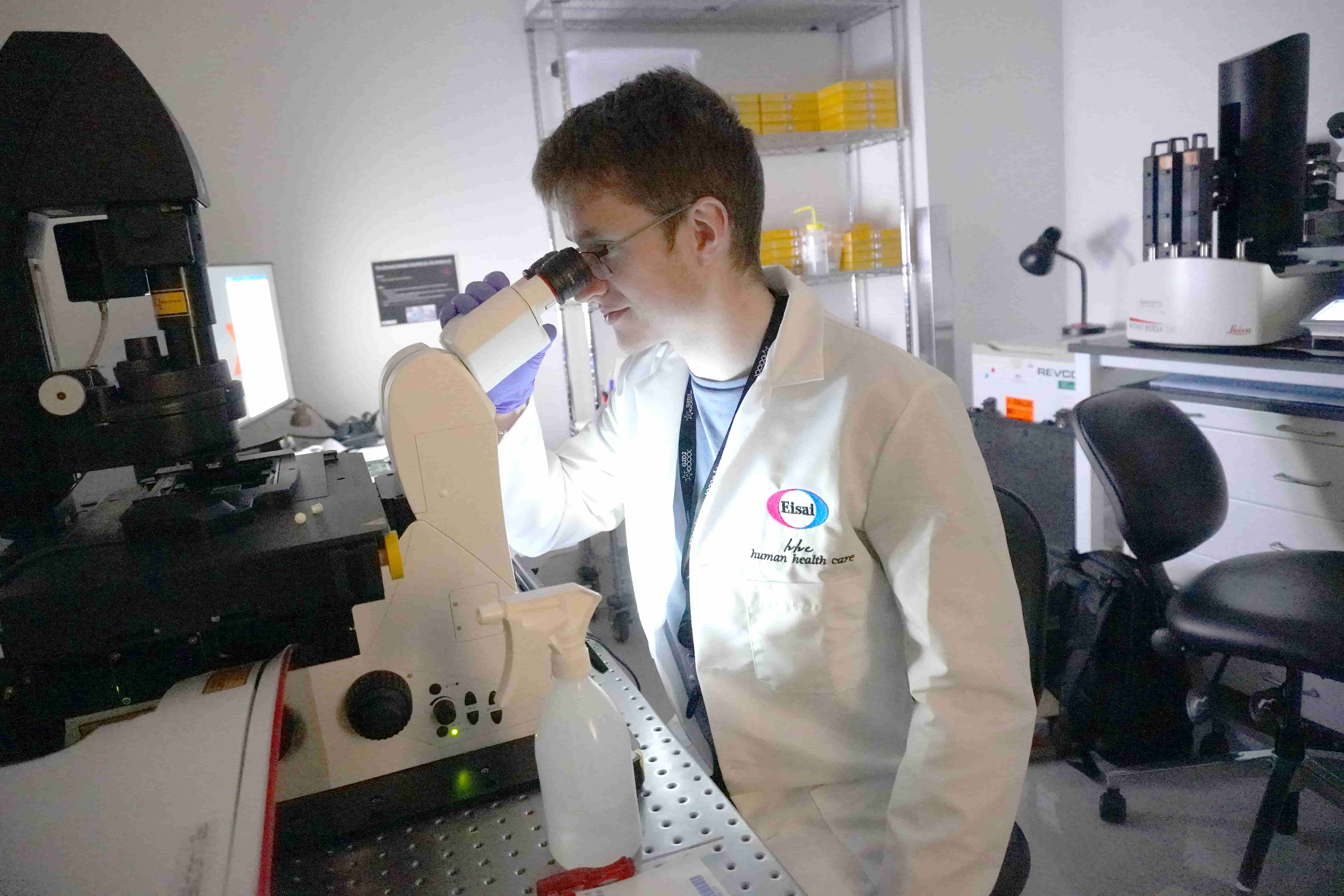 A male scientist wearing an Eisai lab coat looking into a microscope in a lab