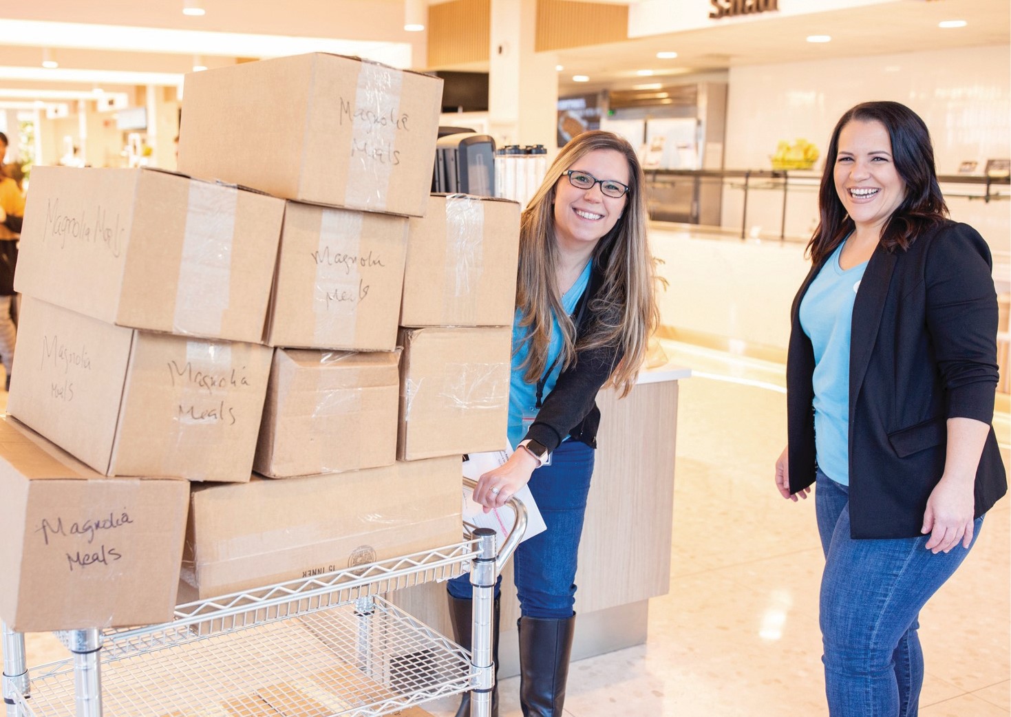 Two smiling women wearing blue shirts in a cafeteria push a cart stacked with cardboard boxes labeled Magnolia Meals.