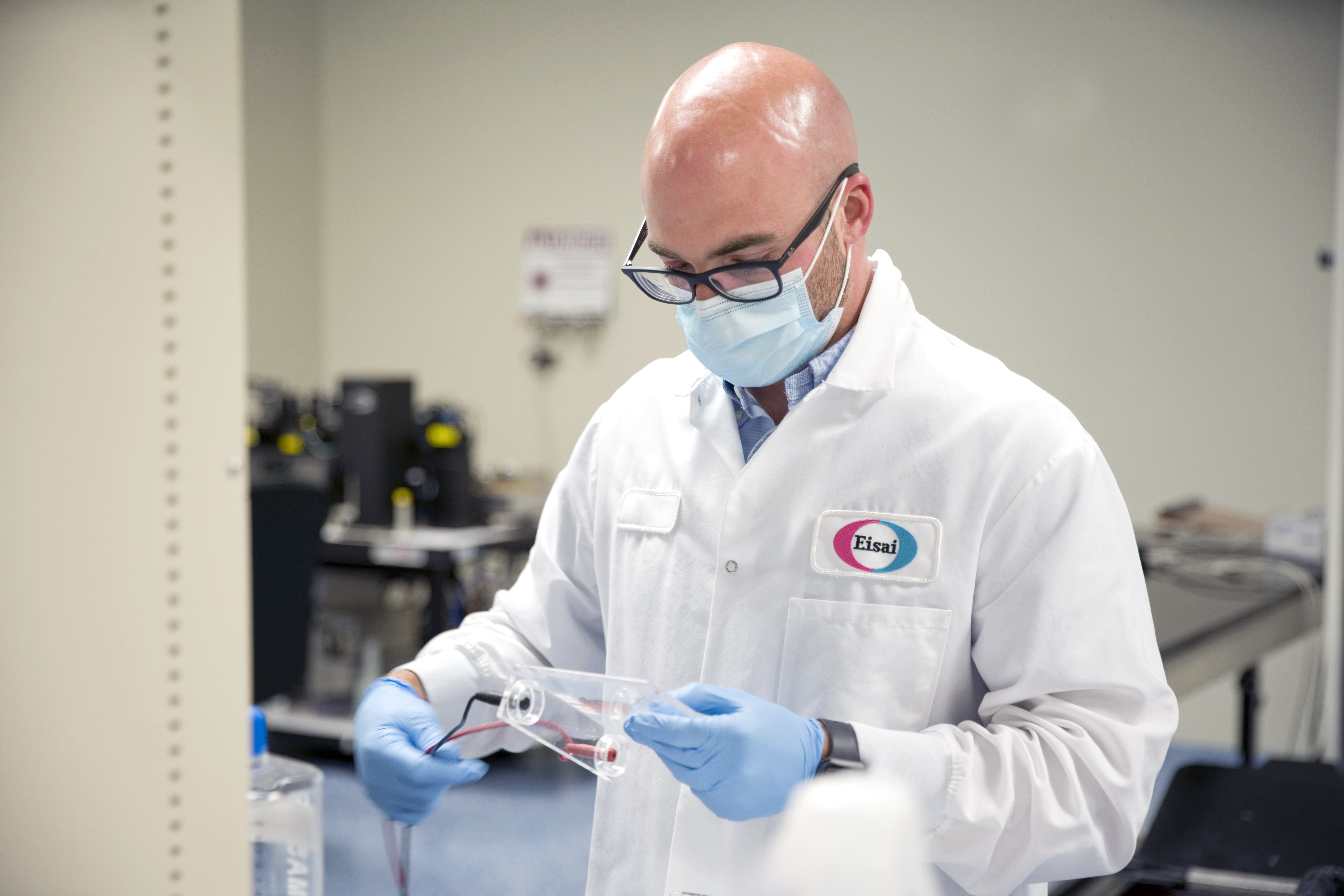 A scientist wearing a lab coat with the Eisai logo, blue gloves, and a blue face mask holds laboratory equipment.