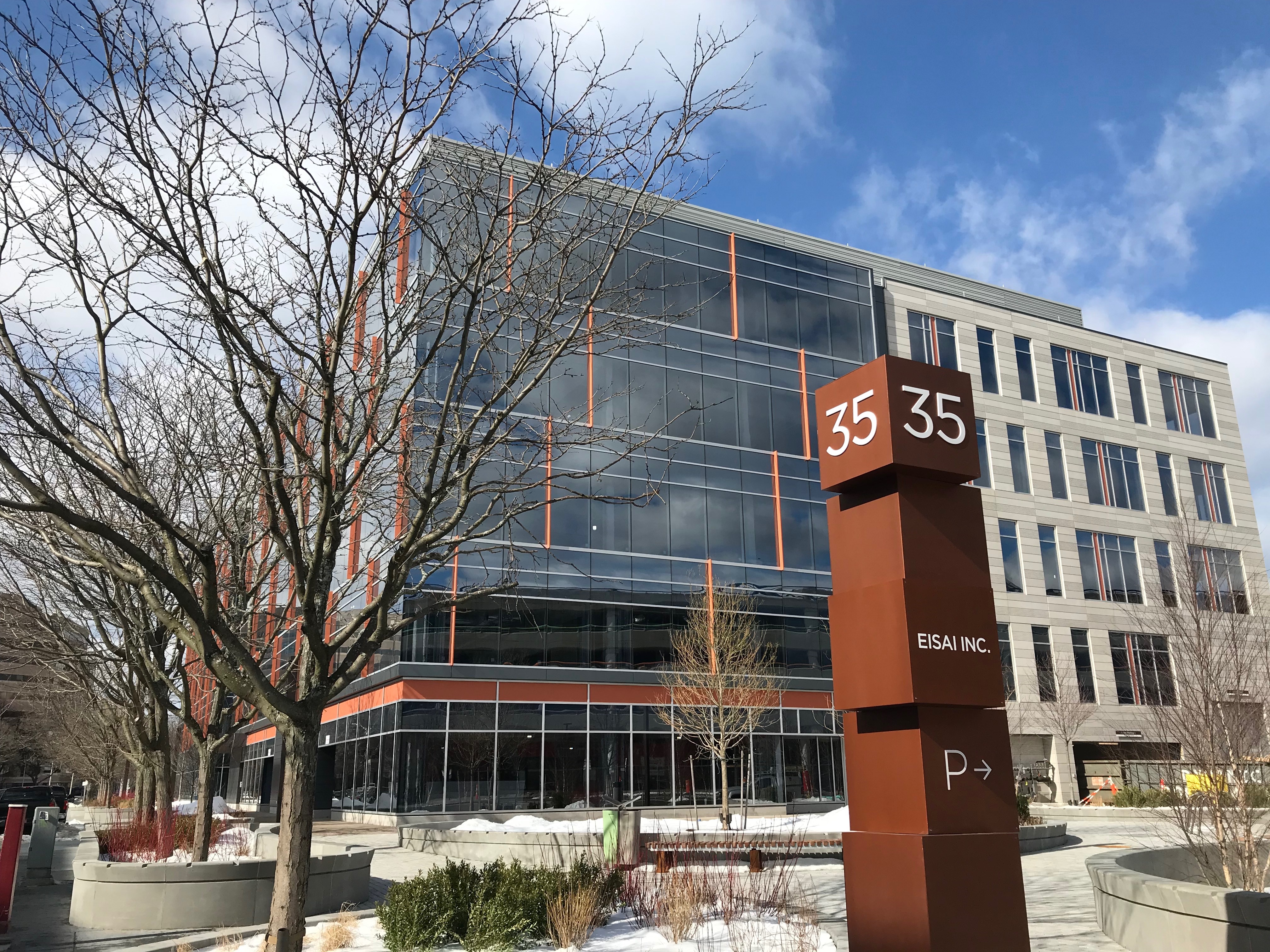 A street-view of a corporate Eisai building with glass exterior with trees and small piles of snow in the foreground.