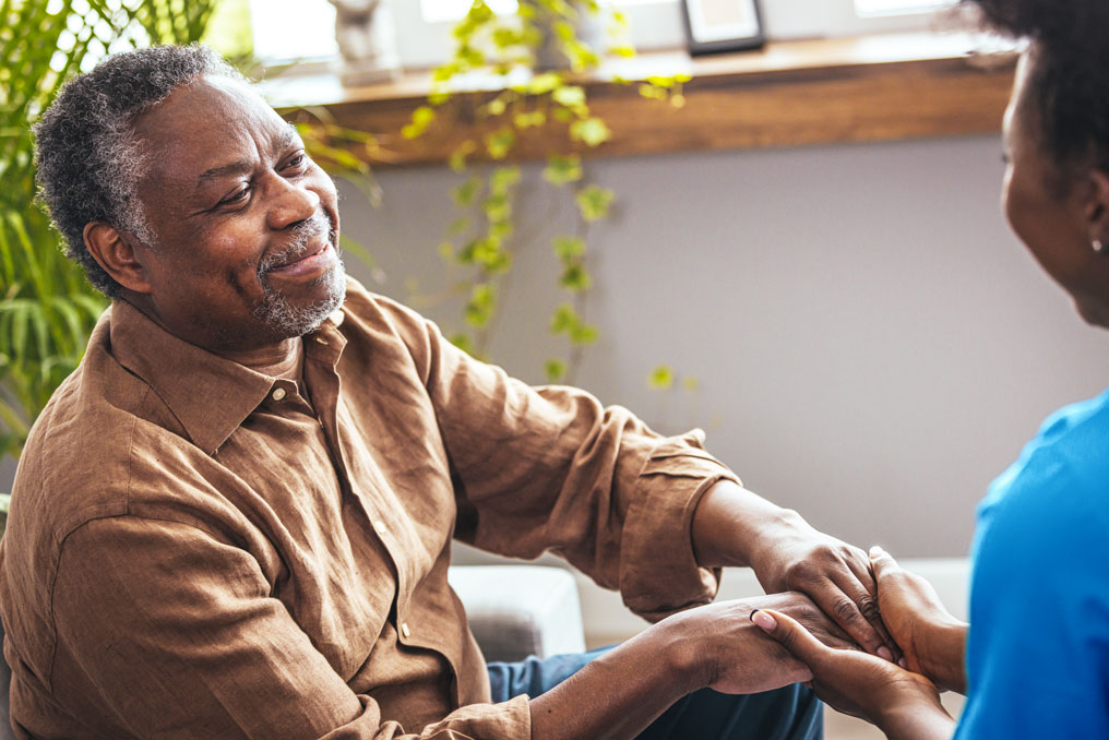 An older man sitting in a sunny room, smiling and facing a woman who is holding his hands.