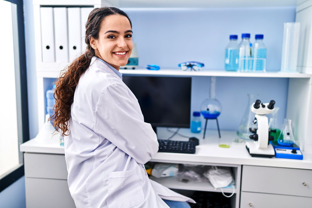 A smilling woman in a medical office setting, wearing a lab coat and sitting at a computer.