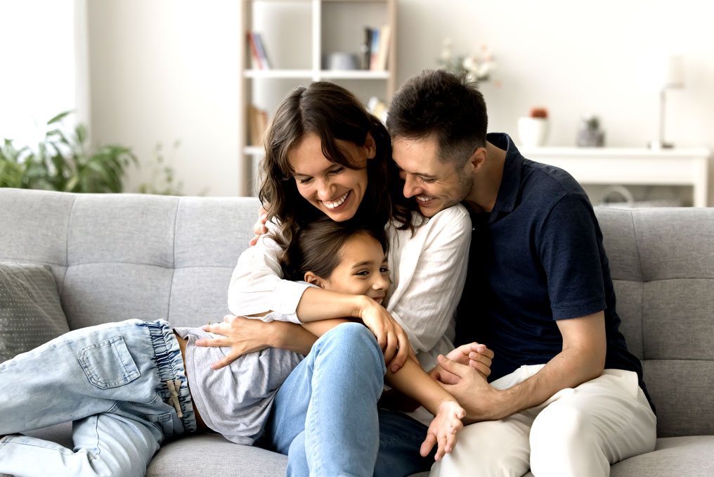 A mom and a dad hugging a child on a grey couch in a sunny room. All are smiling.
